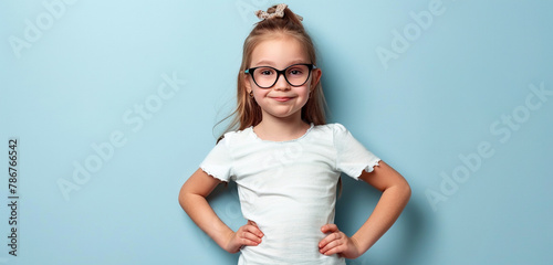 In front of a light blue background, a young girl strikes a playful pose with hands on hips, her glasses adding a hint of maturity to her spirited demeanor