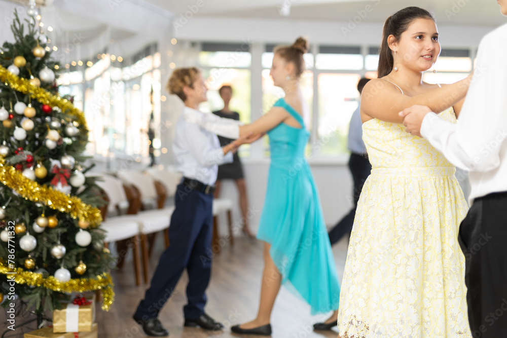Positive teenage girl in festive dress dancing elegant slow ballroom dance with boy partner during New Year party in spacious college hall..