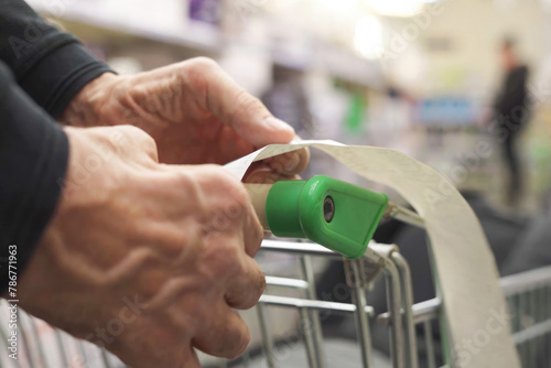 The hands of an unrecognizable older man hold a sales receipt next to a grocery cart in a supermarket.