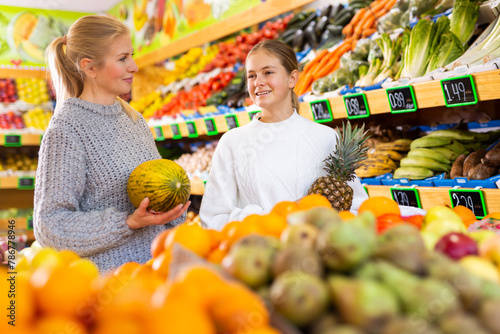 Smiling teenage girl choosing ripe fruits and vegetables, helping her mother to shop in supermarket..