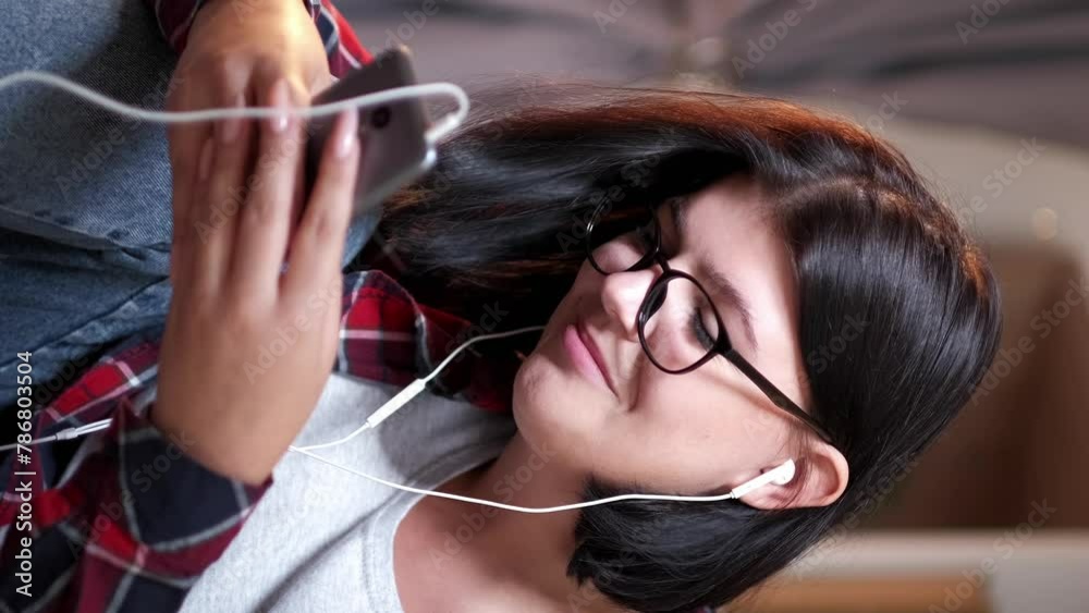 Vertical video. Phone chatting. Gadget leisure. Happy millennial woman typing message on smartphone listening song in earphones home light interior.
