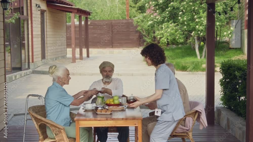 Full shot of smiling female nurse in blue uniform carrying pot of tea for elderly Caucasian people sitting outdoors at retirement home, putting on table, inviting to eat, patting on shoulder, leaving Full shot of smiling female nurse in blue uniform carrying pot of tea for elderly Caucasian people sitting outdoors at retirement home, putting on table, inviting to eat, patting on shoulder, leaving
