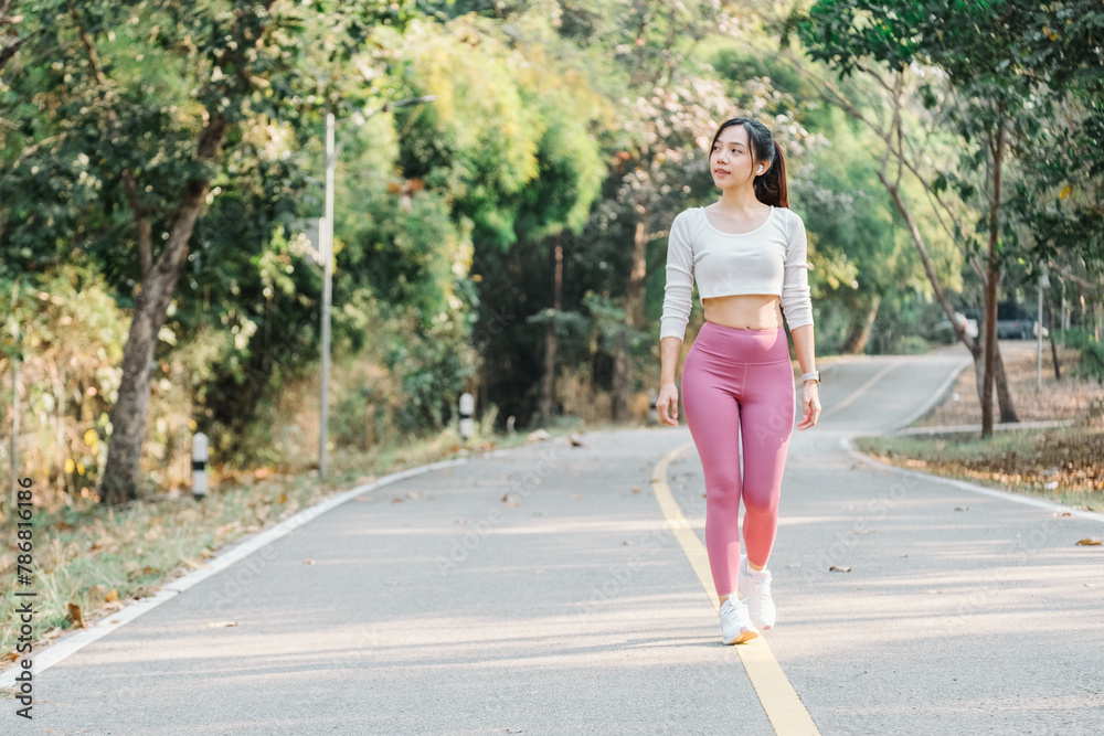Woman in a white cropped top and pink leggings walking on a park path with green trees on the sides.