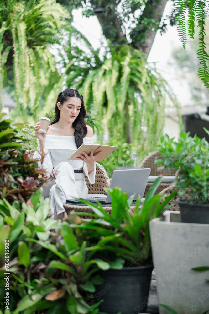 Freelancer deeply engrossed in reading her notes enjoys working outdoors, with a coffee in hand and a laptop nearby in a tranquil garden.