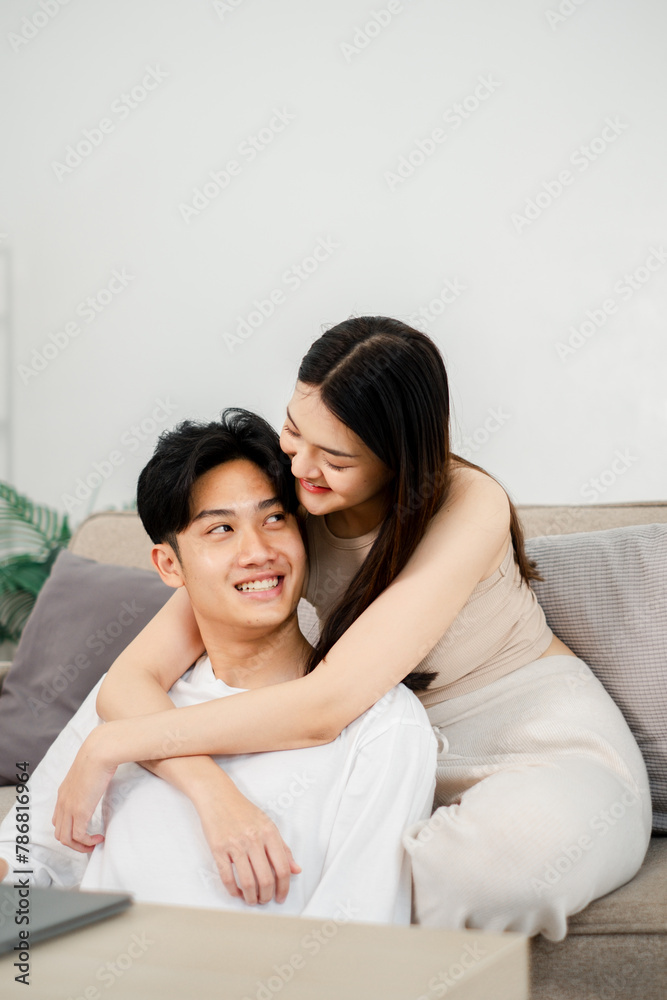 Woman in a beige dress gently embraces a smiling man in a white t-shirt from behind on a gray sofa, creating an intimate moment between them in a modern living room setting. Woman in a beige dress gently embraces a smiling man in a white t-shirt from behind on a gray sofa, creating an intimate moment between them in a modern living room setting.