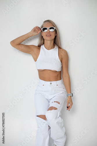 Beautiful slender blonde woman in a white top in white ripped jeans and sunglasses posing on a white background