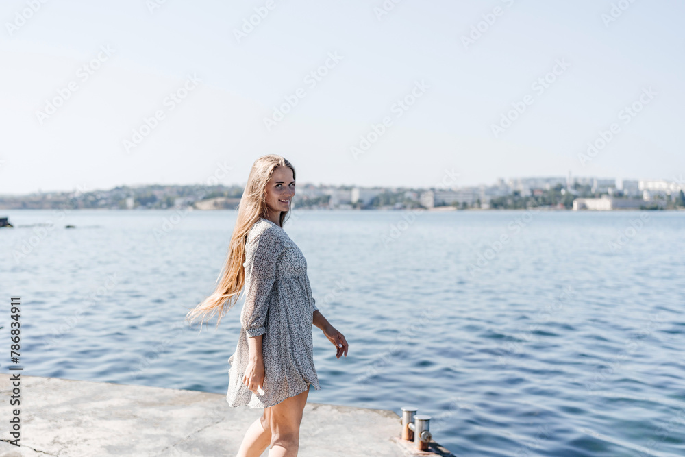 A woman is walking on the beach wearing a dress. The water is calm and the sky is clear.