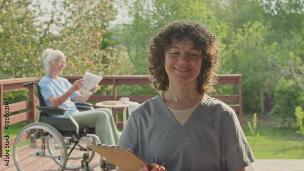 Medium portrait shot of young Caucasian woman in nurse uniform, holding clipboard posing in garden of retirement home, smiling for camera, elderly patient in wheelchair reading newspaper in background Medium portrait shot of young Caucasian woman in nurse uniform, holding clipboard posing in garden of retirement home, smiling for camera, elderly patient in wheelchair reading newspaper in background