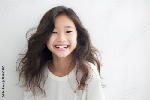 A beautiful Asian woman with long straight hair  whose skin is tan is wearing an all white shirt, and smiling with a face which is full of happiness in a white room. Generative AI. © Surachetsh