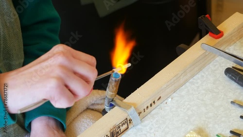 Artisan heats a white rod with a blowtorch, hands in focus, against a wood and metal workbench backdrop. Capturing the essence of craftsmanship and glass bead making photo