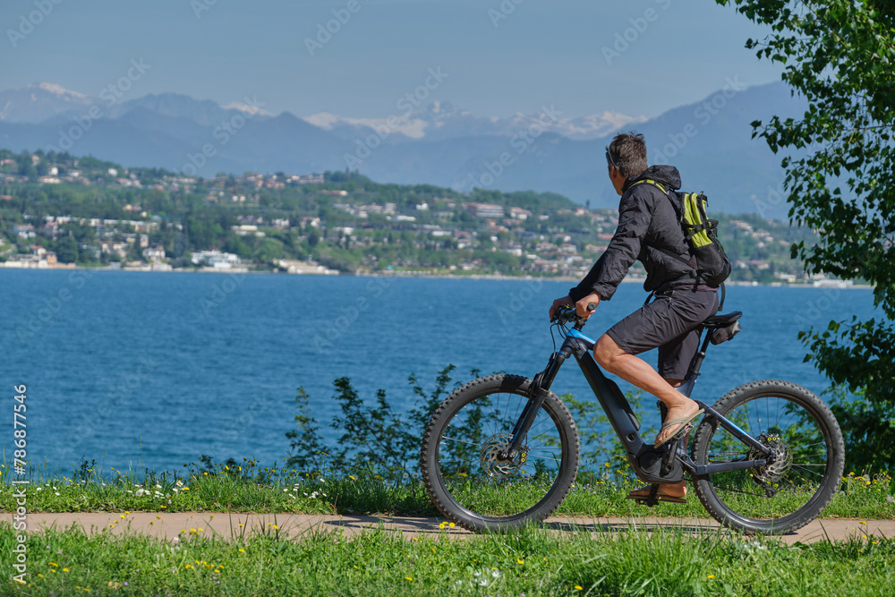 A man in a black jacket and shorts with a backpack on an electric mountain bike in motion in the background Lake Garda, mountain peaks in the snow.