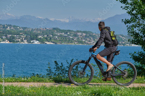 A man in a black jacket and shorts with a backpack on an electric mountain bike in motion in the background Lake Garda, mountain peaks in the snow.