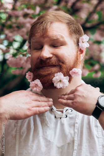 Portrait of a man with a red beard with pink flowers in it. Gender equality. Spring came