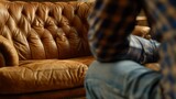 A man in a plaid shirt sits pensively on a classic brown leather couch alone