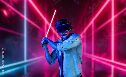Gamer holding laser sword and playing action game while wearing VR glasses. Caucasian man using visual reality headset while standing and surrounding by neon castle. Innovation technology. Deviation.