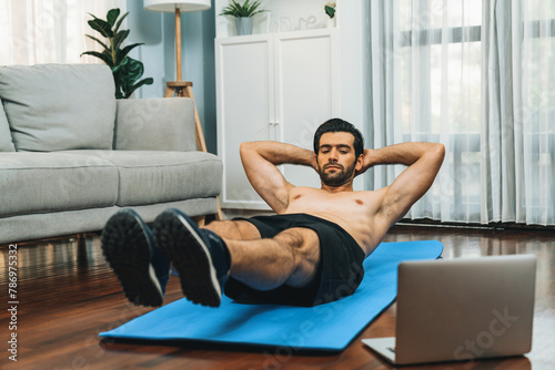 Athletic and sporty man doing crunch on fitness mat while follow online home workout exercise instruction for fit physique and healthy sport lifestyle at home. Online gaiety home exercise video.