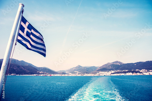 National flag of Greece on a ferry on the background of Greek island landscapes and the trace of the ferry on the water of the sea.