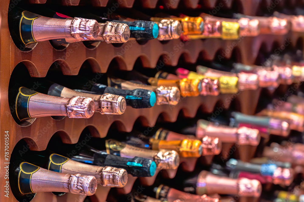 Champagne or cava bottles of different colors lined up and stacked on wooden shelves of a wine rack in a luxury collectible liquor store.