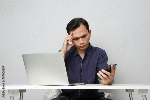 pensive confused asian man holding mobile phone whiile sitting in front of laptop computer. isolated background photo