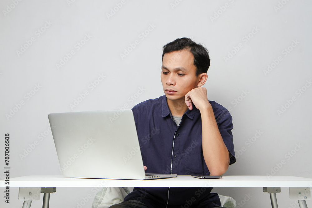 pensive confused asian man holding mobile phone whiile sitting in front of laptop computer. isolated background pensive confused asian man holding mobile phone whiile sitting in front of laptop computer. isolated background