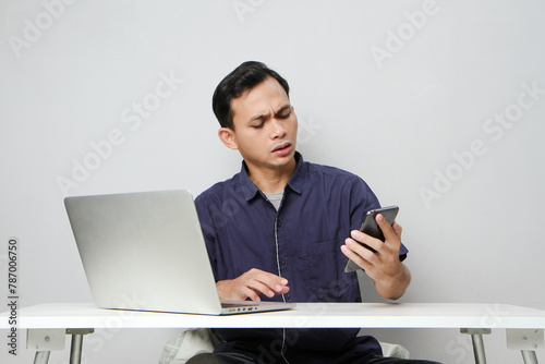 pensive confused asian man holding mobile phone whiile sitting in front of laptop computer. isolated background photo