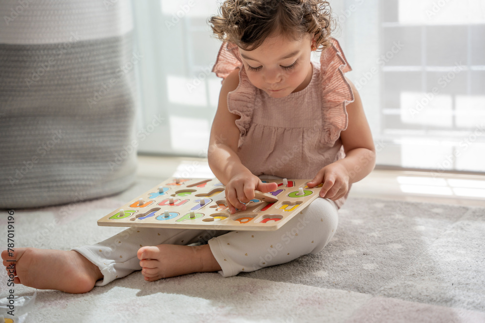 horizontal image of a little girl with curly hair playing sitting on the carpet in her room with a wooden alphabet, Montessori material. concept development and learning in the childhood stage. horizontal image of a little girl with curly hair playing sitting on the carpet in her room with a wooden alphabet, Montessori material. concept development and learning in the childhood stage.