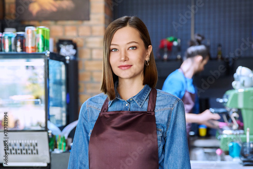 Young woman service worker in apron looking at camera in restaurant, coffee shop