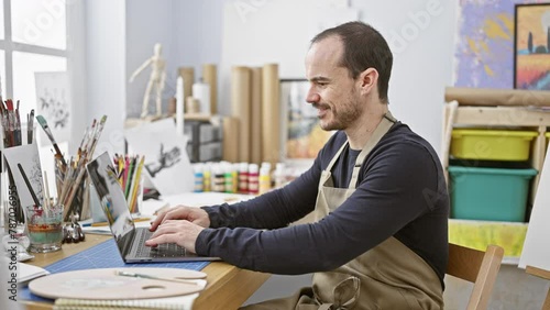 A focused man with a beard and ear piercing works in an art studio, using a laptop surrounded by brushes and paint. photo