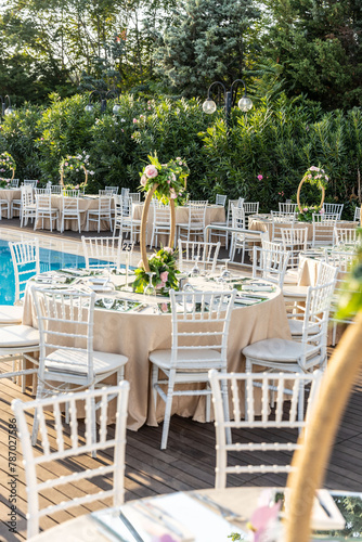 Elegant Outdoor Wedding Reception with Draped Canopy, Floral Centerpieces, and White Chairs by the Poolside Overlooking a Waterfront Venue