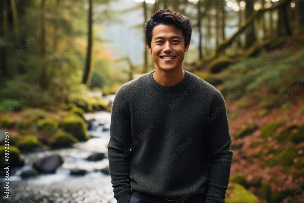 Portrait of a grinning asian man in his 20s dressed in a warm wool sweater over tranquil forest stream