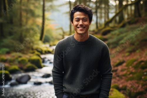 Portrait of a grinning asian man in his 20s dressed in a warm wool sweater over tranquil forest stream