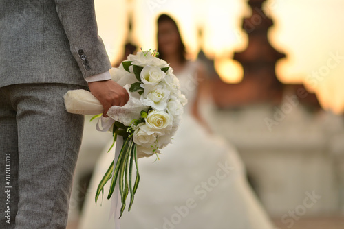 In the wedding ceremony,the groom holding white bouquet waiting for his bride. Both of them stand in front of beutiful Thaistyle building during romantic sunset period. photo