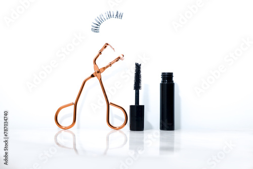 A show off of a fake eyelashes mid-air, it's tweezer and a small mascara place in a white background
