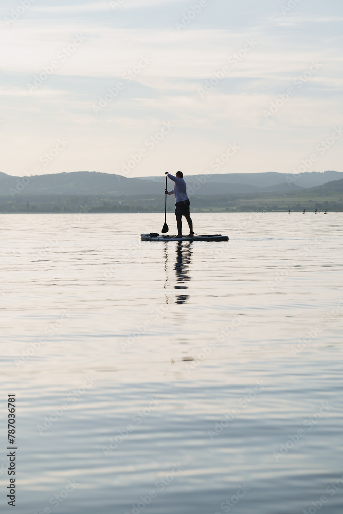Silhouette of a man paddling on a stand up paddle boarder or SUP on the lake. Holding a paddle. Landscape in the background. Active lifestyle.