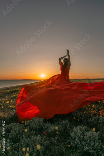 A woman in a red dress is standing in a field with the sun setting behind her. She is reaching up with her arms outstretched, as if she is trying to catch the sun. The scene is serene and peaceful.