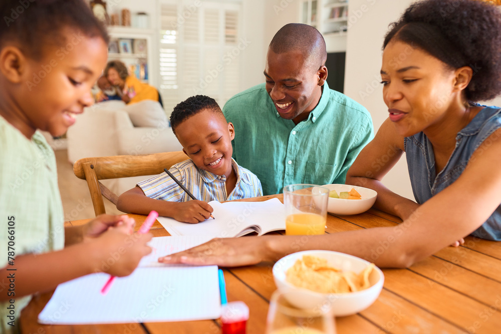 Family Indoors At Home Helping Children With Homework With Grandparents In Background Family Indoors At Home Helping Children With Homework With Grandparents In Background