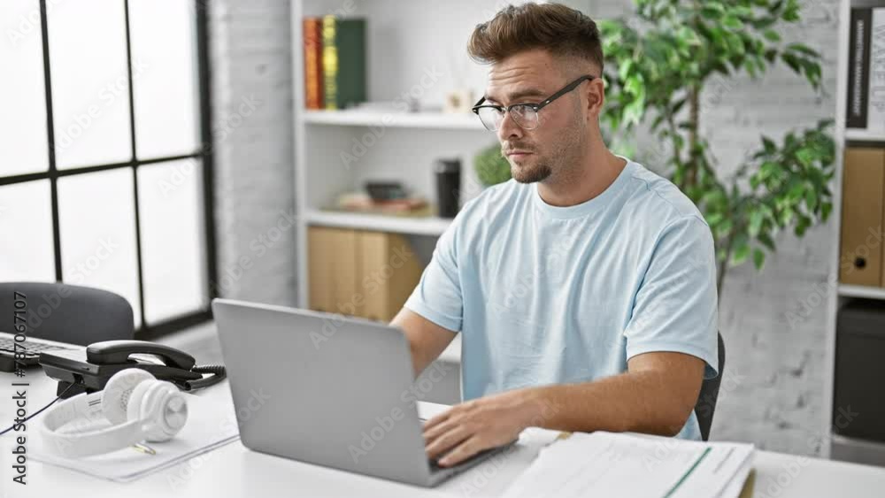 A thoughtful young man with glasses works on laptop in a modern office, evoking professionalism and concentration. A thoughtful young man with glasses works on laptop in a modern office, evoking professionalism and concentration.