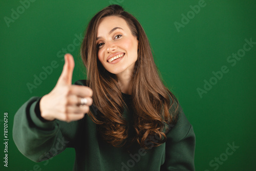 Young pretty woman smiling joyfully and looking happy, feeling carefree and positive with thumb up against green background. Girl wear green pullover. 