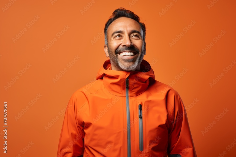 Portrait of a joyful indian man in his 40s wearing a windproof softshell on soft orange background