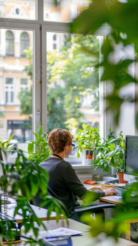 a woman sits at a desk working in front of a window with plants.