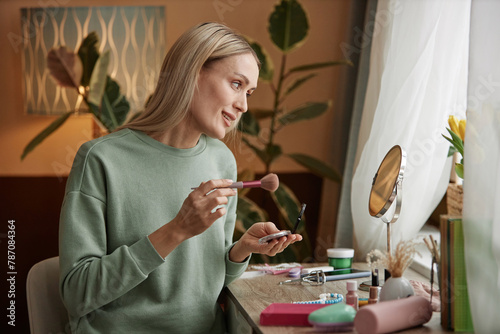 Side view portrait of blonde adult woman doing makeup at home sitting at vanity table by window copy space