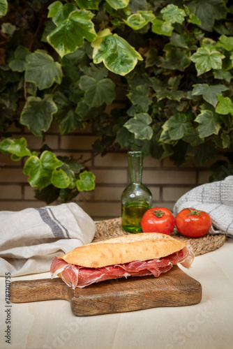 Delicioso bocadillo de jamón serrano con tomate natural y aceite de oliva sobre tabla de madera con fondo con hojas y ladrillo.
