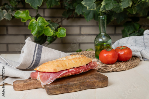 Delicioso bocadillo de jamón serrano con tomate natural y aceite de oliva sobre tabla de madera con fondo con hojas y ladrillo.