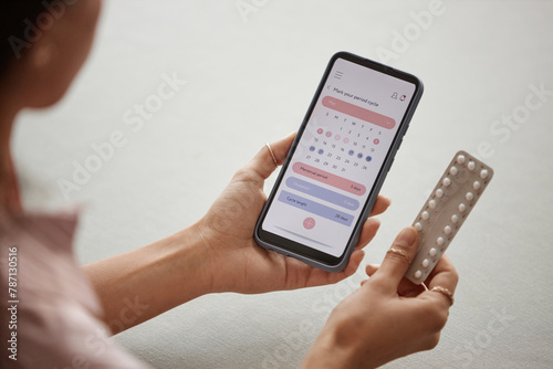 Over shoulder closeup of young woman holding smartphone with calendar app on screen and pack of birth control pills