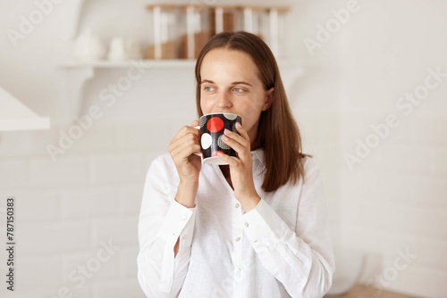 Positive Dark Haired Female Wearing White Casual Style T Shirt Drinking Coffee Tea Morning Posing With Kitchen Set Background Looking Away With Dreamy Expression