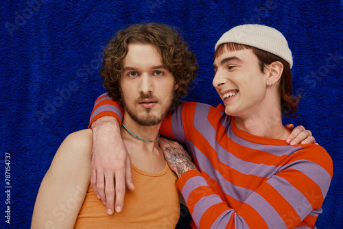 two joyful good looking gay men in vibrant clothes posing on dark blue backdrop, pride month photo