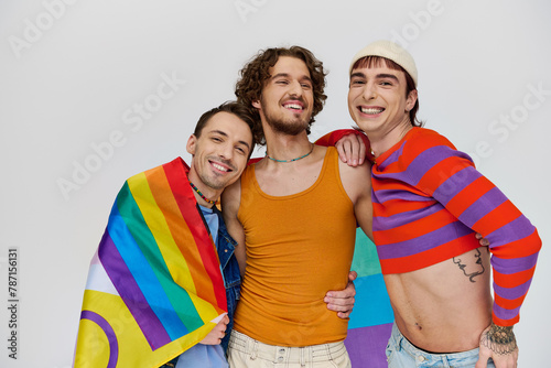 three joyous stylish gay men in cozy clothing posing actively with rainbow flag on gray backdrop photo