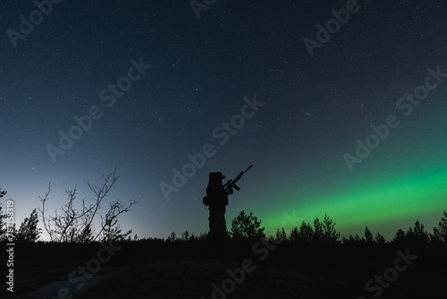 Soldier with a night vision device and a rifle with a suppressor in the forest against the background of the starry sky and northern lights.