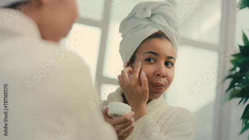 Happy woman in head towel applying undereye cream in front of mirror, skincare photo