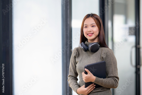 Free Download Happy young Asian woman holding a tablet with headphones around her neck in a bright, modern office. - GFXLoad